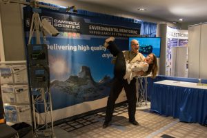 Photograph of two people at a Campbell Scientific exhibit stand at the CMOS congress. Man is shown holding woman in the air. Both are laughing.