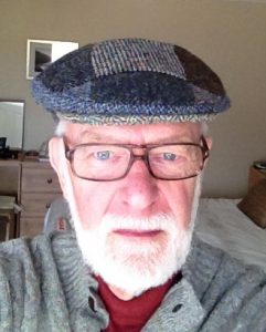 Photograph shows an older man with a white beard and glasses wearing a tweed hat.