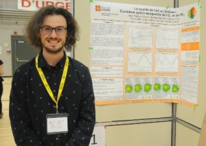 Photo shows a smiling Jean-Philippe Gilbert standing beside a scientific poster. He is in his early 20's, chin length dark hair, beard and moustache, with glasses.