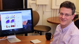 Photo shows a smiling Paul Kushner, caucasian male, clean shaven, 40's, dark hair and glasses, sitting adjacent to a computer screen.