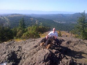 Photograph shows a caucasian man wearing sunglasses kneeling at the top of a rocky mountain, with two alsatian dogs at his feet. Tree-filled slopes appear in the background.