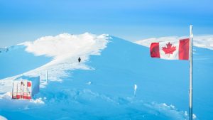 Photo shows a Canada flag waving in the foreground beside a building, and a person in the distance walking along a snowy ridge at the PEARL ridge lab.