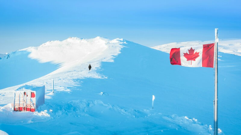 Photo shows a Canada flag waving in the foreground beside a building, and a person in the distance walking along a snowy ridge at the PEARL ridge lab at Eureka.