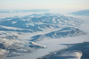 Photo shows an aerial long-distance view of a snow and ice landscape, mountains and a river channel.