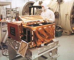 The MOPITT instrument shown here as a square metallic box with an open lid, and a man in a white clean suit overseeing it, undergoing test at the University of Toronto around 1995. (Photo credit: COMDEV)