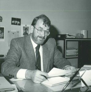 Black and white photo of Phil Merilees. Photo shows a caucasian man with eyeglasses and a beard, sitting at a desk. Circa 1975.