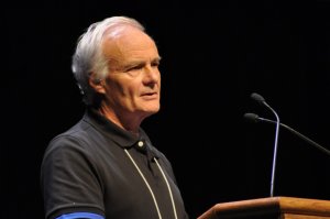 Photo of a middle aged man, grey hair, standing at a podium, Robie MacDonald of CMOS