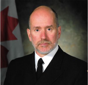 Headshot of a younger-middle aged Caucasian male with grey stubble beard, black suit and tie with Canadian flag in the background.