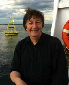 Smiling caucasian middle aged man with dark hair, standing on a boat at sea, leaning on the rail, with a yellow buoy in the background