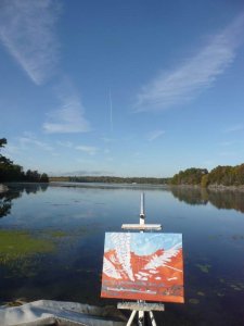 landscape painting in progress on canvas on an easel by the lake. Painting is red, with white marks and blue sky..
