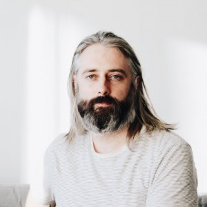 Headshot of man in white sweater and long grey hair with a brown beard