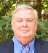 Smiling Caucasian man with blue collared shirt and blazer, standing in front of a pine tree