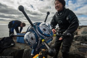 A person in a wetsuit who has just exited the water with a large scientific instrument for ocean monitoring in the foreground.