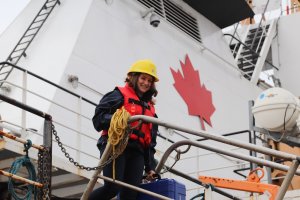 A person in an yellow helmet and orange life vest smiling and standing on a reserach vessel with a red maple leaf painted on the vessel