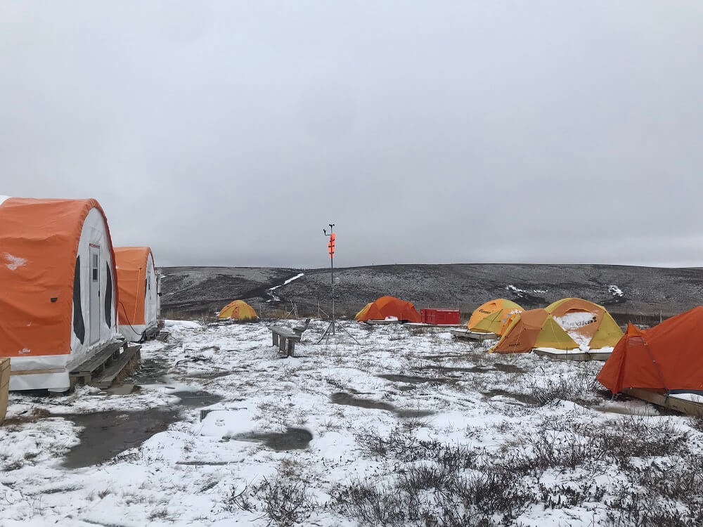 Several orange camping tents and two large prospector tents on the snowy tundra. Some scientific instruments set up between the tents. 