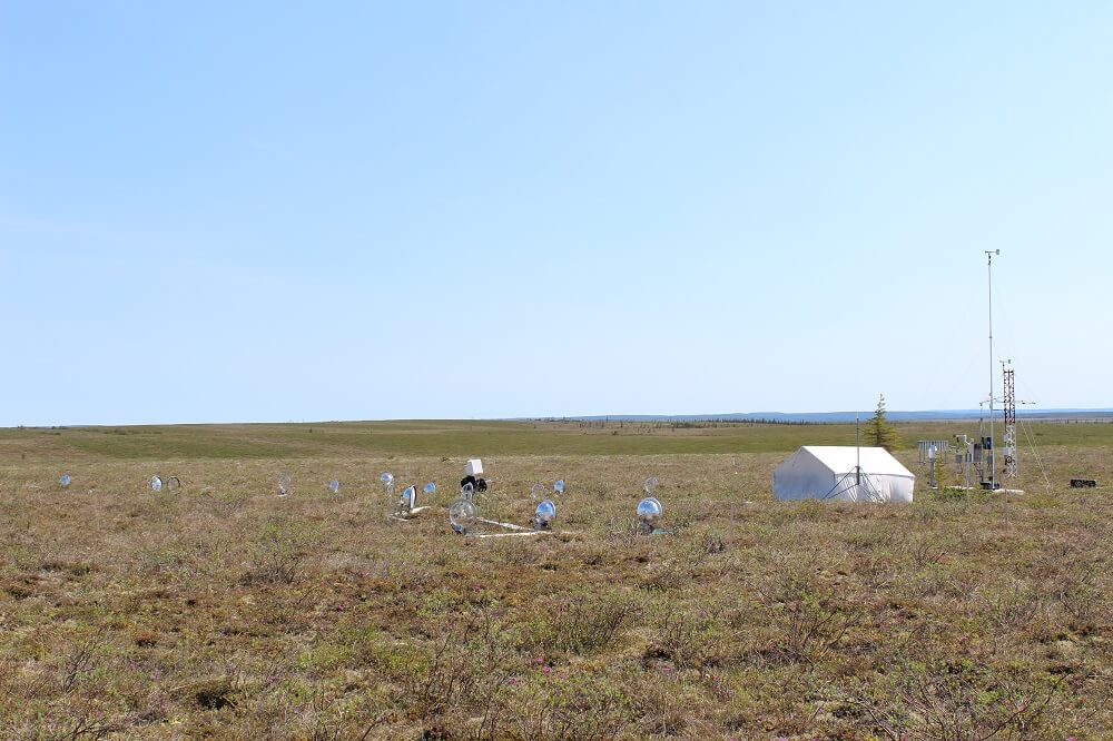 A landscape view of the tundra with a large white tent and scientific instruments on the landscape. 