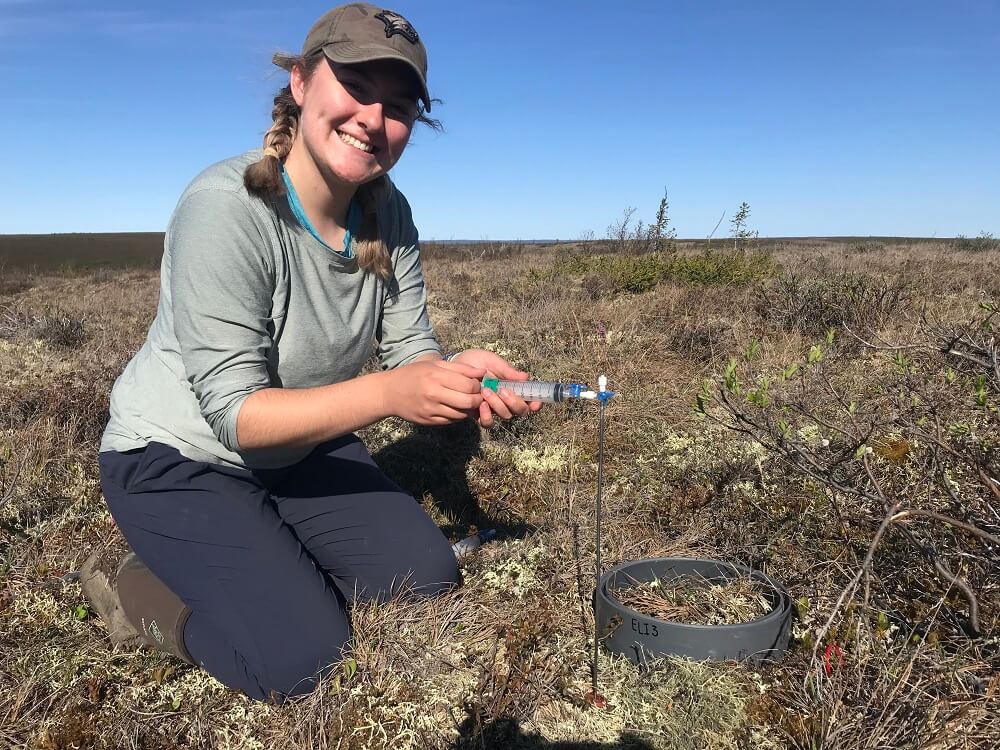 A young woman kneeling on the tundra smiling and holding up a scientific syringe.