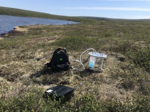 A backpack, a laptop and a transparent chamber sitting on the tundra next to a lake.