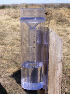 A transparent rain gauge 1/3 filled with water on a woodend post in a field of dry grass