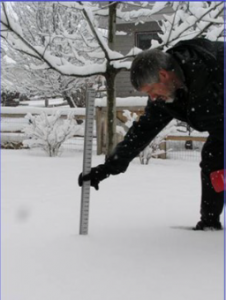 A man with grey hair wearing a black winter coat bending over in the snow with a ruler placed into the snow on the ground