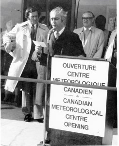 An old black and white photo of a man in a trench coat standing beside a podium with sign indicating the opening of the Canadian Meteorological centre. The man is talking into a microphone and holding up a cue card,