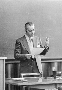 An old black and white photograph of a man in a suit standing in front of a desk looking at a paper and talking with an animated hand