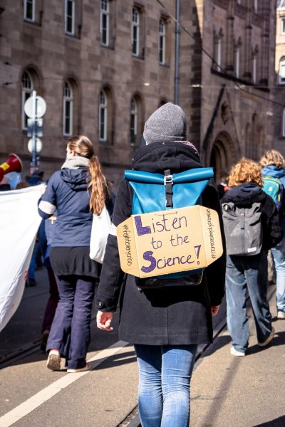 The back of a person in winter clothing walking at a protest with a sign on their back reading Listen to the Science