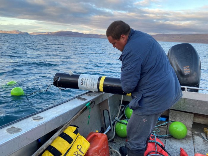 A man standing at the edge of a boat with his back to the camera deploying a scientific instrument into the water