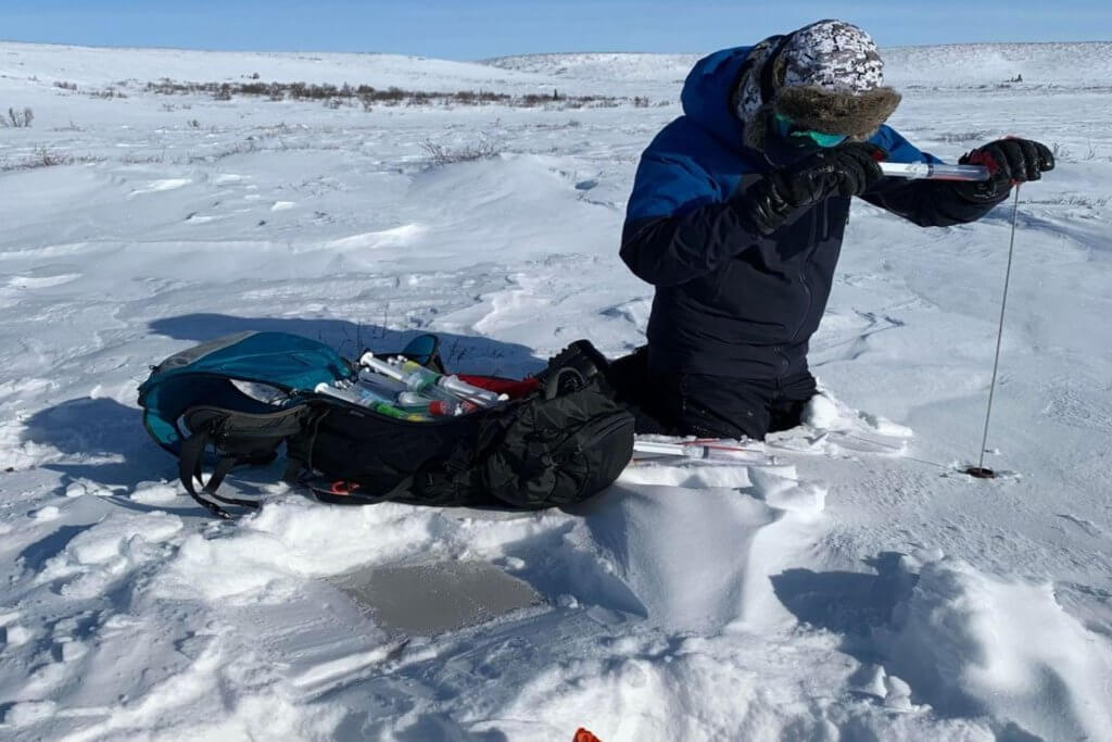 A person dressed in heavy winter gear kneeling on the snow with the tundra landscape in the balckground. The person is holding a metal pole which they are inserting into the snow. Beside the person is a large backpack with several syringes laid on top.