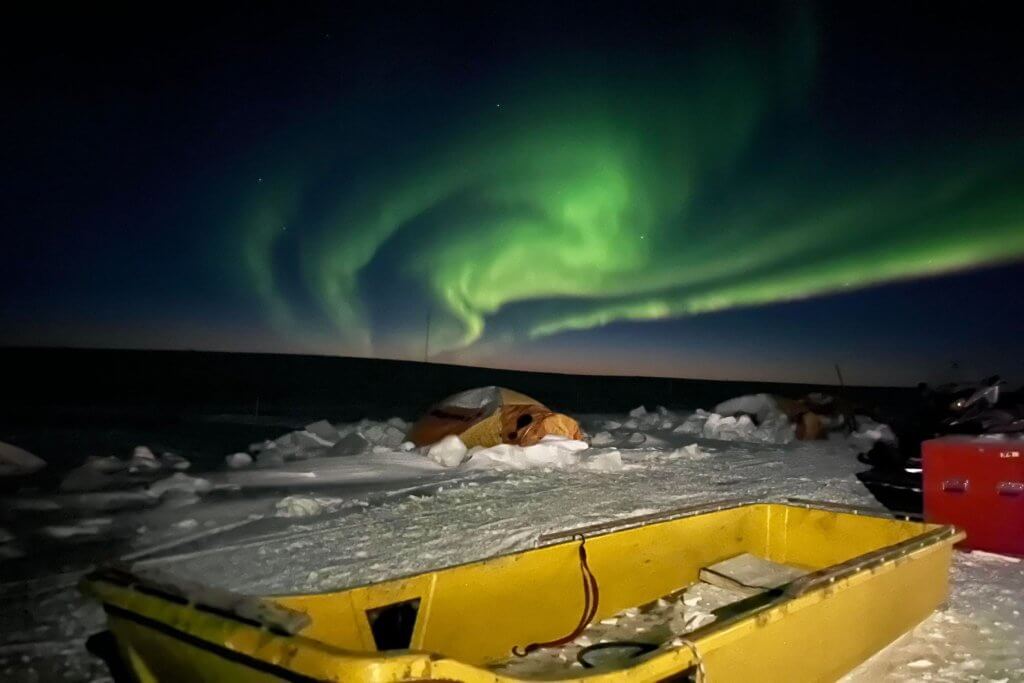 Landscape photo of the northern lights with a yellow sled and and orange tent in the foreground