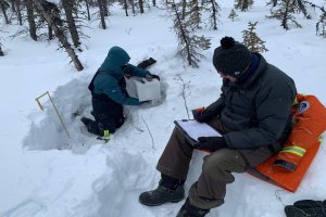 One person sitting in a pit dug into the snow holding a white box with some electronic sensors on top. Another person sitting beside the snow pit writing on a clipboard