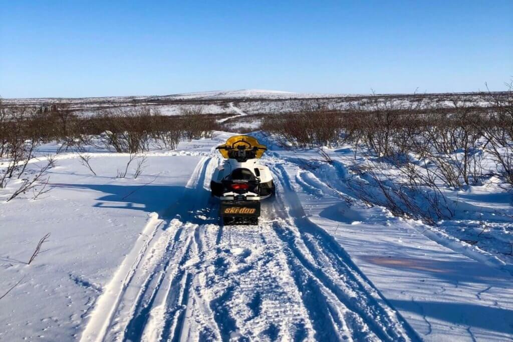 A snowmobile parked on a snowmobile track between some tundra bushes on the snow on a sunny day