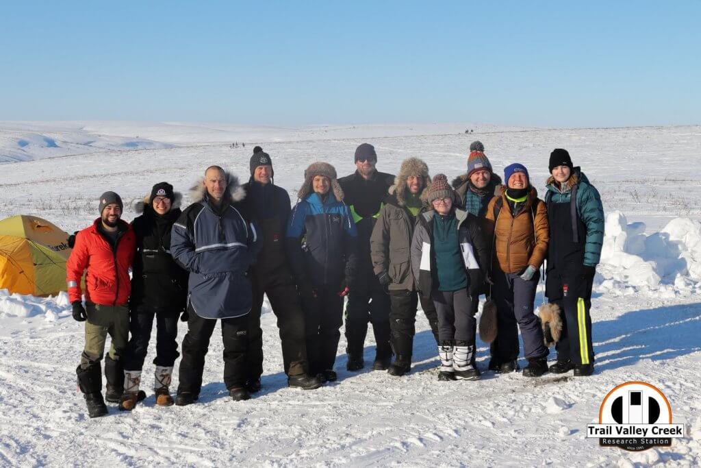 Eleven people standing in the snowy tundra and smiling for the camera. They are dressed in heavy winter gear and it is a sunny day with blue sky